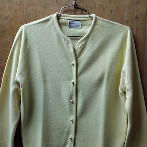 May include: Pale yellow cardigan sweater with a round neckline and long sleeves. The sweater has a row of pearl-like buttons down the front. The label reads "Tarralan". The sweater is hanging on a metal hanger.
