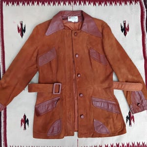 Può includere: Una giacca vintage in camoscio marrone con chiusura a bottoni, cintura e inserti in pelle sul colletto, tasche e polsini. La giacca ha un design classico e probabilmente risale agli anni '70. La giacca è esposta su un tessuto fantasia.