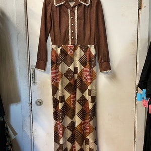 May include: A long brown dress with a patchwork skirt. The skirt is made of different fabrics in brown, white, and red. The dress has a white lace collar and buttons down the front.