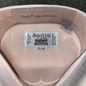 Pode incluir: Uma camisa rosa para homem com uma etiqueta branca que diz "Single Needle Tailoring by martini" e "65% Poliéster 35% Algodão". A etiqueta também diz "Machine Wash Warm" e "Made in Korea AN 21512". A camisa tem uma etiqueta de tamanho que diz "15-35".