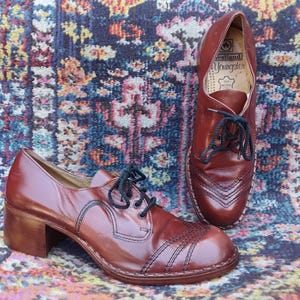 May include: A pair of vintage brown leather shoes with black laces. One shoe is upright, showcasing the interior label. The other shoe is angled, revealing a block heel and decorative stitching. The shoes are on a patterned rug.