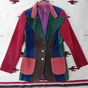 May include: A colorful corduroy blazer with a patchwork design. The jacket features panels of red, green, blue, and brown, with a purple interior and two front pockets. Gold buttons secure the front. The blazer is laid on a patterned rug.