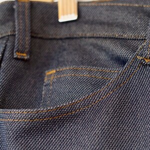 May include: Close-up of a dark blue denim pocket with orange stitching. The denim has a textured weave.