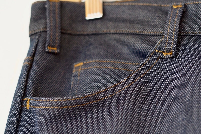 May include: Close-up of a dark blue denim pocket with orange stitching. The denim has a textured weave.