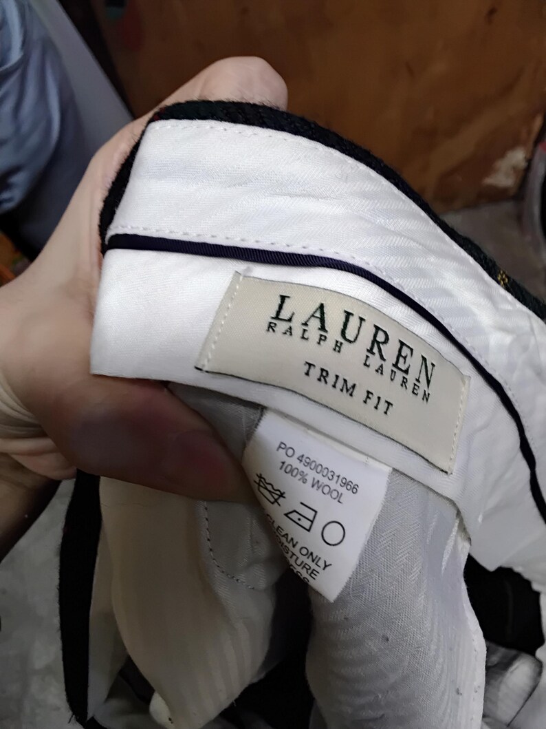 May include: Close-up of the interior of a pair of black wool trousers. The white lining is visible, along with a label that reads "LAUREN RALPH LAUREN TRIM FIT" and a care label. The trousers are made of 100% wool.