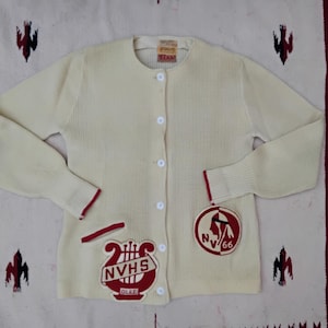 May include: Cream-colored vintage cardigan sweater with red trim and patches. Features a round neckline, button-up front, and ribbed cuffs. Patches include a lyre with "NVHS GLEE" and a Native American head with "NV 66".