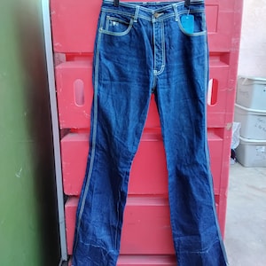 May include: Dark blue denim jeans with a flared leg, hanging on a black hanger. The jeans have white stitching along the seams and pockets. A blue tag hangs from the front pocket. The jeans are displayed against a red background.