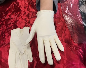 May include: Two pairs of white leather gloves on a red satin surface. One pair is laid flat, while the other pair is held by a hand.