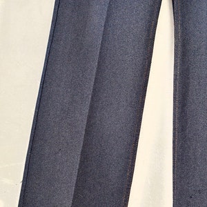 May include: Close-up of the dark blue denim fabric of a pair of jeans. The fabric has a subtle texture and is sewn with a light brown thread.