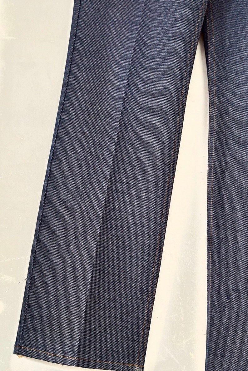 May include: Close-up of the dark blue denim fabric of a pair of jeans. The fabric has a subtle texture and is sewn with a light brown thread.
