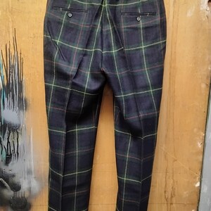 May include: Dark navy blue plaid trousers with green, red, and yellow accents. The pants are hanging on a black hanger, showcasing the back pockets and button details. The plaid pattern is a classic design.