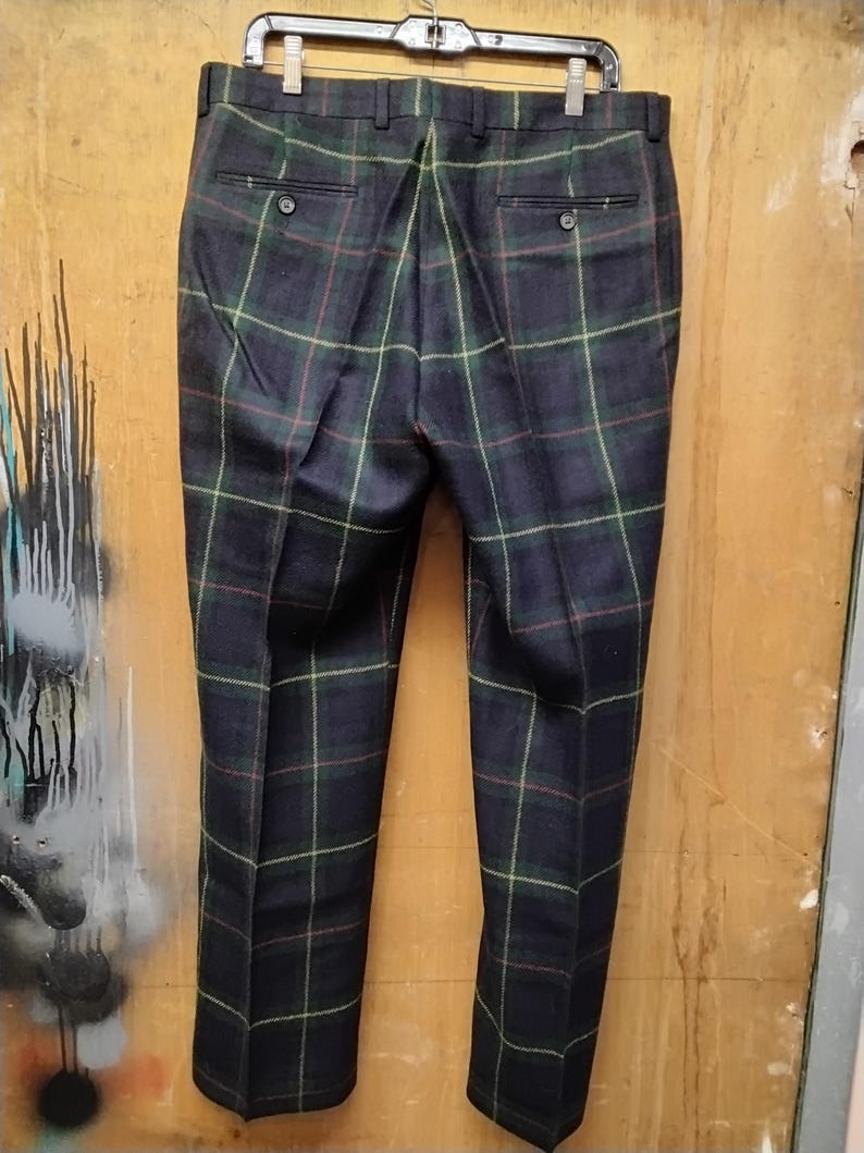 May include: Dark navy blue plaid trousers with green, red, and yellow accents. The pants are hanging on a black hanger, showcasing the back pockets and button details. The plaid pattern is a classic design.