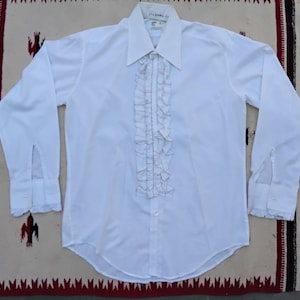 Pode incluir: Uma camisa branca de manga comprida com uma frente com babados e punhos abotoados. A camisa tem uma gola clássica e está colocada sobre um tecido estampado.