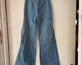 Puede incluir: Un par de pantalones de campana de mezclilla azul claro colgados de una percha. Los pantalones tienen cintura alta y pierna ancha.