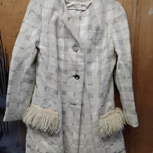 May include: A vintage, light-colored coat with a checkered pattern. The coat features a round collar, two buttons, and fringed pocket details. The coat is hanging on a metal hanger.
