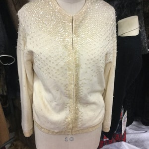 May include: A white cardigan sweater with a beaded design. The cardigan has a zip closure and is on a white mannequin. The label on the mannequin reads "Royal Form".