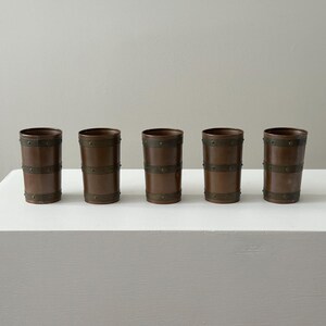May include: Five copper-toned drinking glasses with a barrel design. Each glass has dark metal bands around the top, middle, and bottom, secured with small rivets. The cylindrical glasses stand on a white surface. The glasses are approximately 12 cm tall.