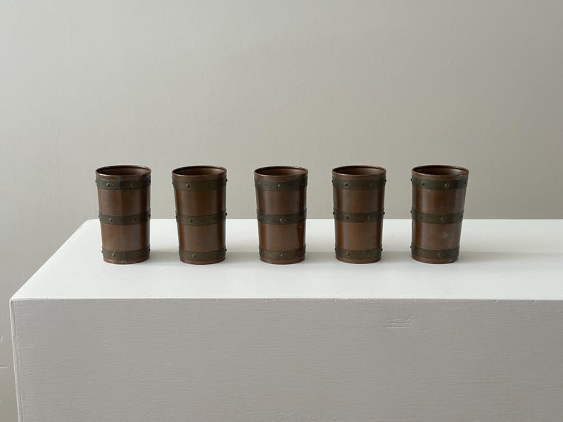 May include: Five copper-toned drinking glasses with a barrel design. Each glass has dark metal bands around the top, middle, and bottom, secured with small rivets. The cylindrical glasses stand on a white surface. The glasses are approximately 12 cm tall.