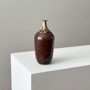 May include: A handcrafted, brown ceramic vase with a textured surface. The vase has a narrow neck and a wider body, with a light brown and gold rim. The vase is displayed on a white surface.