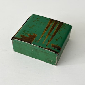 May include: A small, rectangular green metal box with a hinged lid. The box has a distressed finish with brown and gold geometric designs on the top. The box is likely vintage or antique.
