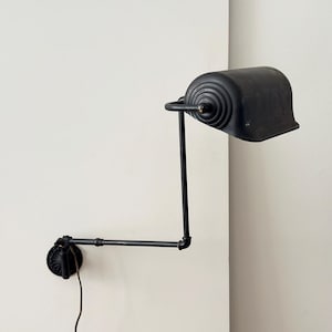 May include: A black, wall-mounted lamp with an adjustable arm and a conical shade. The lamp is made of metal and has a vintage industrial design. The shade is approximately 6 inches wide.