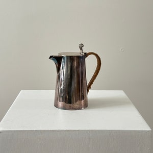 May include: A silver pitcher with a brown woven handle and a silver lid. The pitcher is on a white surface.