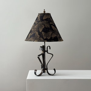 May include: A decorative table lamp with a black and gold butterfly pattern lampshade. The lamp has a dark metal base with a unique, artistic design. The lamp is placed on a white surface.