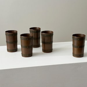 May include: Five copper-coloured drinking glasses with a barrel design. Each glass has two horizontal bands of metal rivets. The glasses are cylindrical and stand upright on a white surface.