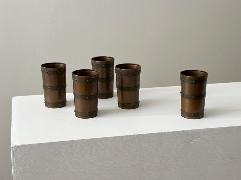 May include: Five copper-coloured drinking glasses with a barrel design. Each glass has two horizontal bands of metal rivets. The glasses are cylindrical and stand upright on a white surface.
