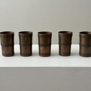 May include: Five copper-coloured drinking glasses with dark metal bands. The glasses are cylindrical with a slightly flared top. The bands are evenly spaced around the body of each glass. They are displayed on a white surface.