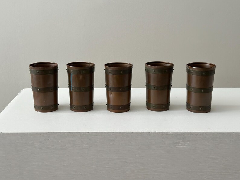 May include: Five copper-coloured drinking glasses with dark metal bands. The glasses are cylindrical with a slightly flared top. The bands are evenly spaced around the body of each glass. They are displayed on a white surface.