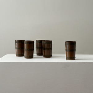 May include: Five brown barrel-shaped drinking glasses with metal bands. The glasses are arranged on a white surface against a neutral background. The glasses have a rustic, vintage aesthetic.