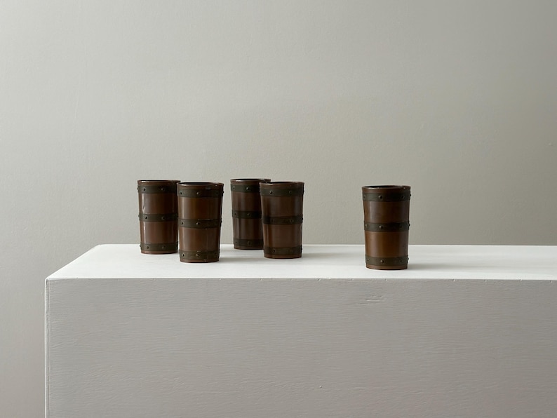 May include: Five brown barrel-shaped drinking glasses with metal bands. The glasses are arranged on a white surface against a neutral background. The glasses have a rustic, vintage aesthetic.