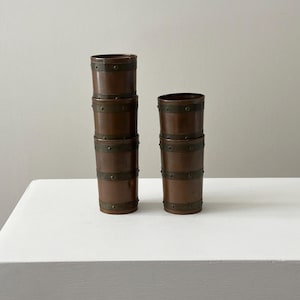 May include: Four brown wooden cups with metal bands are stacked on a white surface. Two stacks of cups are visible, one with three cups and the other with two. The cups have a barrel-like design.