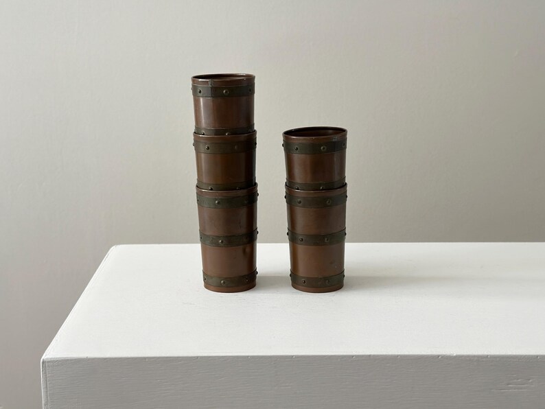 May include: Four brown wooden cups with metal bands are stacked on a white surface. Two stacks of cups are visible, one with three cups and the other with two. The cups have a barrel-like design.