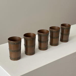 May include: Five copper-coloured tumblers with brass-coloured bands. The cylindrical glasses have a rustic, vintage appearance. The bands are evenly spaced around the body of each glass. The glasses are arranged in a row on a white surface.
