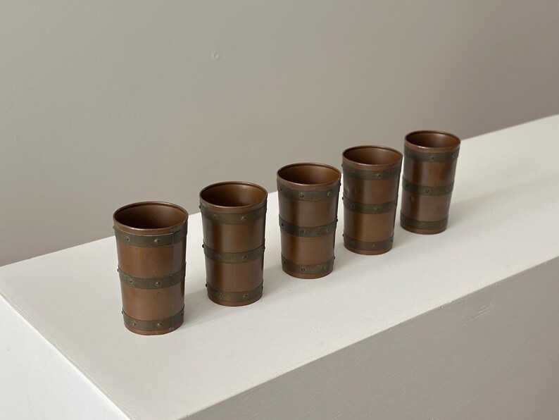 May include: Five copper-coloured tumblers with brass-coloured bands. The cylindrical glasses have a rustic, vintage appearance. The bands are evenly spaced around the body of each glass. The glasses are arranged in a row on a white surface.