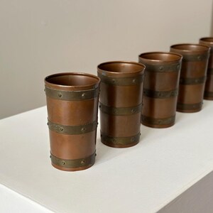 May include: Five copper-coloured metal tumblers with brass-coloured bands. The cylindrical cups are arranged diagonally on a white surface. The metal has a warm, reddish-brown hue, and the bands are riveted.