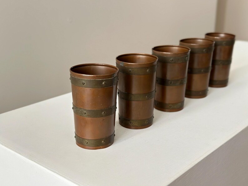 May include: Five copper-coloured metal tumblers with brass-coloured bands. The cylindrical cups are arranged diagonally on a white surface. The metal has a warm, reddish-brown hue, and the bands are riveted.