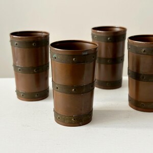 May include: Four copper-coloured tumblers with brass bands. The cylindrical glasses have a vintage aesthetic, with two brass bands encircling each tumbler. The glasses are arranged on a white surface.