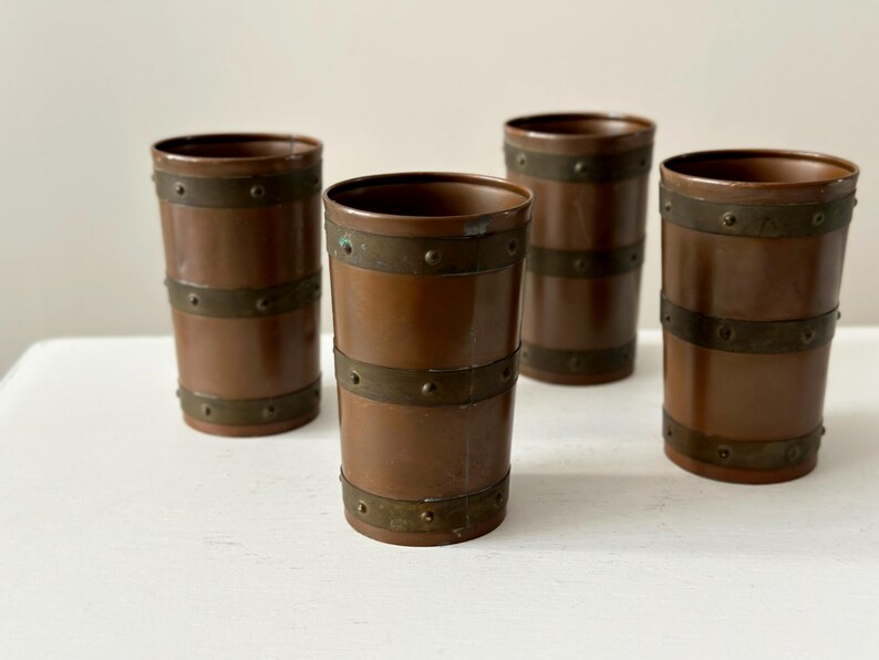 May include: Four copper-coloured tumblers with brass bands. The cylindrical glasses have a vintage aesthetic, with two brass bands encircling each tumbler. The glasses are arranged on a white surface.