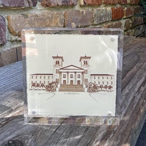 May include: A framed sepia-toned line drawing of a large building with two towers. The building has a central entrance with a set of stairs leading up to it. The drawing is on a white background and is framed in a clear acrylic frame.