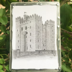 May include: A black and white pencil drawing of a castle on a white background. The drawing is framed in a silver metal frame. The castle has a tall tower with a pointed roof and a smaller tower on the right side. There is a gate in the front of the castle.