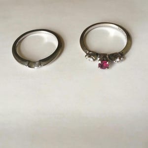 May include: Two silver rings are displayed on a white surface. One ring is a simple band, while the other features a central red gemstone flanked by smaller clear stones. The rings are likely for engagement or fashion.
