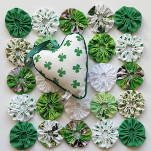 May include: A heart-shaped shamrock-patterned ornament with a green trim and a green ribbon, surrounded by green and white fabric circles. The fabric circles have various patterns, including shamrocks and floral designs. The arrangement is on a white surface.