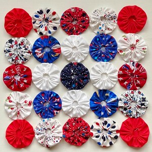 May include: A collection of twenty-five circular fabric yo-yos arranged in a five-by-five grid. The fabric pieces feature various patterns including solid red, white, blue, star prints, and cherry motifs, each with a gathered center hole.