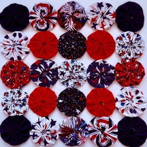 May include: A collection of twenty-five fabric yo-yos arranged in a five-by-five grid. The circular fabric pieces feature various patterns including solid red, solid navy blue, white with blue stars, red and white stripes, and firework motifs.
