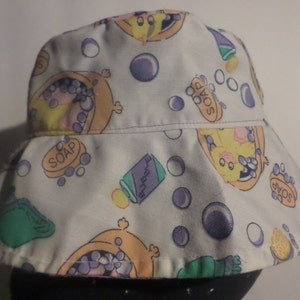 Puede incluir: Sombrero de cubo blanco con un estampado de dibujos animados de un personaje amarillo tomando un baño con jabón y burbujas. El sombrero tiene un borde morado y una corona blanca.