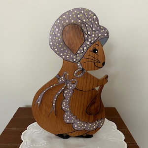 May include: Wooden mouse figurine with a bonnet and apron, painted in shades of brown, purple, and white. The mouse is holding a small bag. The figurine is placed on a white, scalloped doily.