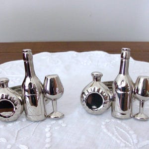 Könnte beinhalten: Zwei Sets silberfarbener Serviettenringe in Form von Weinflaschen und Gläsern. Jedes Set enthält eine Flasche, eine Karaffe und ein Weinglas. Die Serviettenringe sind auf einem weißen, bestickten Stoff ausgestellt.